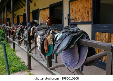 Horse riding saddles hanging on the fence of a stable with horses on background. Close up, copy space for text.
