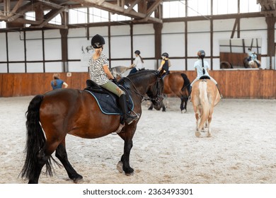 Horse riding school. Little children girls at group training equestrian lessons in indoor ranch horse riding hall. Cute little beginner blond girl kid in helmet sitting on brown horse horseback - Powered by Shutterstock