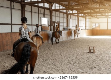 Horse riding school. Little children girls at group training equestrian lessons in indoor ranch horse riding hall. Cute little beginner blond girl kid in helmet sitting on brown horse horseback - Powered by Shutterstock