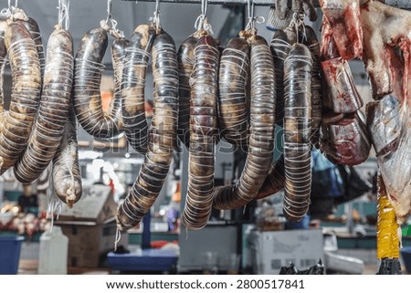 Horse sausage (kazy). Long, dried, segmented carcasses hang on metal hooks. Market stalls display raw, dried, and prepared meat products. The image conveys traditional style.
