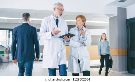 In the Hospital, Busy Doctors Talk, Using Tablet Computer While Walking Through the Building. In the Background Patient Talks with Receptionist. New Modern Fully Functional Medical Facility.