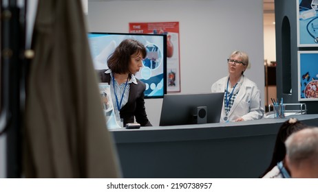 Hospital Employee Working On Appointment Service At Reception Desk, Giving Administrative Information To Doctor And Patients In Waiting Room. Medical Clinic Lobby With Help Counter.