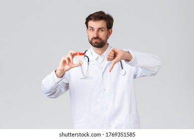 Hospital, healthcare workers, covid-19 treatment concept. Displeased serious doctor in white scrubs and stethoscope, showing medication pills and thumbs-down, dont recommend using drugs