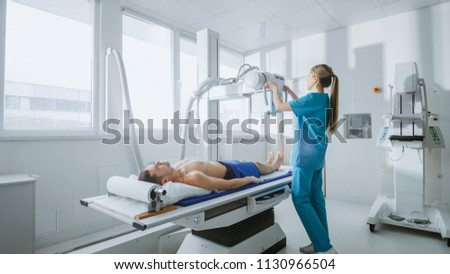 In the Hospital, Low Angle Shot of Man Lying on a Bed, Female Technician adjusts X-Ray Machine. Scanning for Fractures, Broken Limbs, Injuries, Cancer or Tumor. Modern Hospital