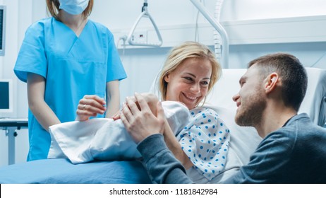 In The Hospital Midwife Gives Newborn Baby To A Mother To Hold, Supportive Father Lovingly Hugging Baby And Wife. Happy Family In The Modern Delivery Ward.