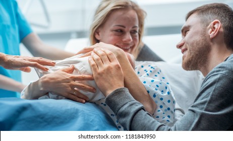 In The Hospital Midwife Gives Newborn Baby To A Mother To Hold, Supportive Father Lovingly Hugging Baby And Wife. Happy Family In The Modern Delivery Ward.