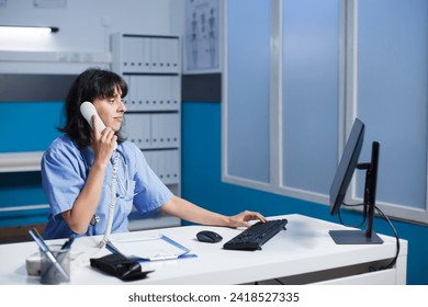 In the hospital office, a female medical professional takes calls and sets up appointments. Nurse utilizes a telephone at a desk in a contemporary clinic while focusing on a pc monitor. - Powered by Shutterstock