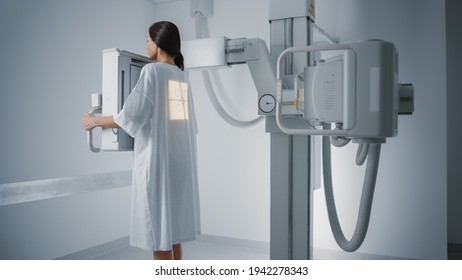 Hospital Radiology Room: Beautiful Multiethnic Woman Standing in Medical Gown in the X-Ray Machine. Adult Female Undergoes Healthcare Exam and is Scanning Chest, Heart, Lungs in Modern Clinic Office.