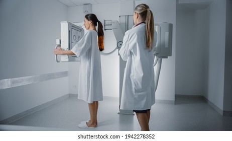 Hospital Radiology Room: Beautiful Multiethnic Woman In Medical Gown Standing Next To X-Ray Machine While Female Doctor Adjusts It. Healthy Patient Undergoes Routine Scanning With The Nurse's Help.