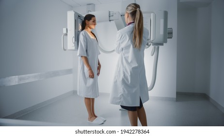 Hospital Radiology Room: Beautiful Multiethnic Woman In Medical Gown Standing Next To X-Ray Machine While Female Doctor Adjusts It. Healthy Patient Undergoes Routine Scanning With The Nurse's Help.