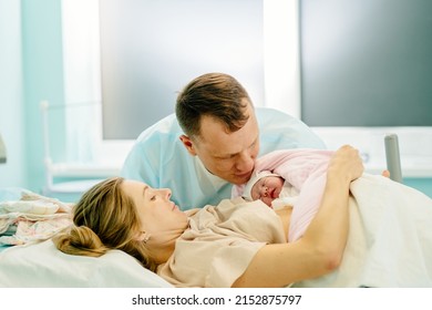 In Hospital Tired Mother Holding Newborn Baby Girl, Supportive Father Lovingly Kissing Baby And Wife. Family In The Delivery Ward.