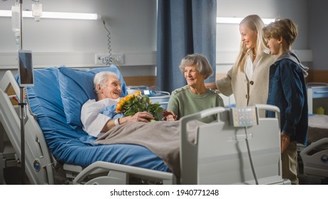 Hospital Ward: Elderly Man Resting in Bed, His Caring Beautiful Wife Beside, Happy Grandson and Daughter Visit, Give Flowers. Family Support Old Man Recovering After Successful Surgery