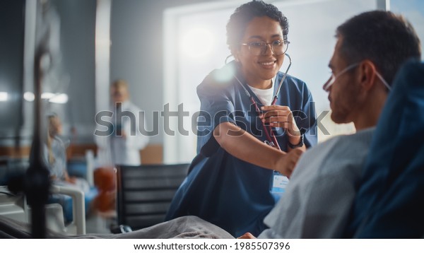 Hospital Ward: Friendly Black Head Nurse Uses
Stethoscope to Listen to Heartbeat and Lungs of Recovering Male
Patient Resting in Bed, Does Checkup. Man Getting well after
Successful Surgery