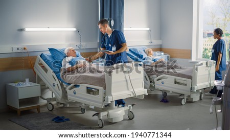 Hospital Ward: Friendly Male Nurse Talks Reassuringly to Elderly Patient Resting in Bed. Doctor or Physician Uses Tablet Computer, Does Checkup, Old Man Fully Recovering after Successful Surgery