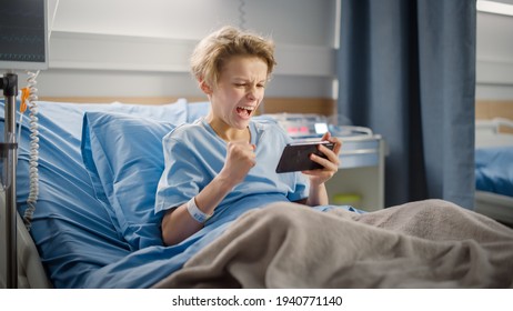 Hospital Ward: Handsome Young Boy Resting in Bed, Uses Smartphone, Wins in Online Video Game on Internet with Mobile Phone. Happy Patient Recovering after Sickness - Powered by Shutterstock