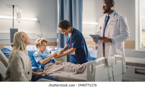Hospital Ward: Handsome Young Boy Resting in Bed with Caring Mother Visits to Support Him, Friendly Doctor Talks, Head Nurse Monitoring Child's Vitals. Happy Smiling Patient Recovering after Sickness