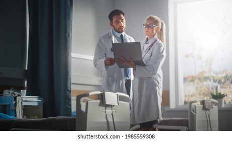 Hospital Ward: Male And Feamle Professional Doctors Hold Laptop Computer, Talk About Patient Medical History, Discuss Health Care Advice, Treatment Plan. Modern Clinic With Advanced Equipment