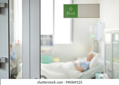 Hospital Ward Sign On Closed Glass Door In Clinic With Blurred Shape Of Senior Patient Lying On Bed Behind It