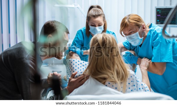 In the Hospital Woman Giving Birth, Husband\
Holds Her Hand in Support, Obstetricians Assisting. Modern Delivery\
Ward with Professional\
Midwives.