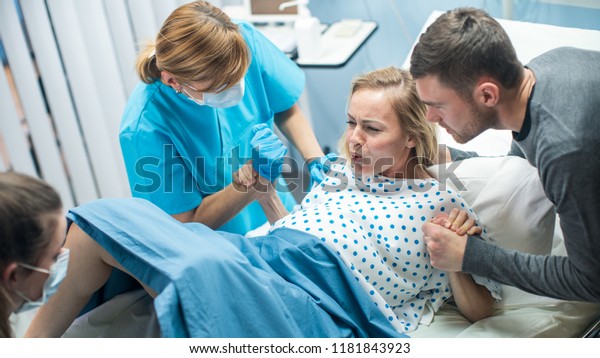 In the\
Hospital, Woman in Labor Gives Birth, Obstetricians and Doctors\
Assist, Her Husband Supports Her by Holding Hand. Modern Maternity/\
Delivery Ward with Professional\
Midwives.