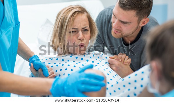 In the Hospital Woman in\
Labor Pushes to Give Birth, Obstetricians Assisting, Husband Holds\
Her Hand for Support. Modern Delivery Ward with Professional\
Midwives.