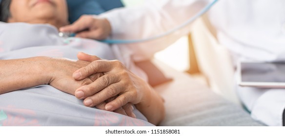 Hospitalized Elderly Patient, Senior Old Aging Woman Laying On Bed With Cardiologist Doctor Or Physician Examining Cardiological Heart Health, Checking Pulse In Hospital Clinic Exam Room