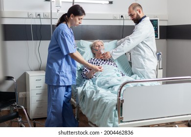 Hospitalized Senior Woman Inhale And Exhale Through Oxygen Mask Laying In Hospital Bed. Medical Nurse Using Stethoscope Listening Patient Heart.