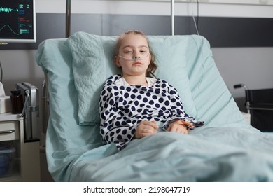 Hospitalized Sick Little Girl Resting In Children Healthcare Clinic Facility Patient Bed. Under Treatment Ill Kid Laying In Hospital Pediatric Ward While Looking At Camera.