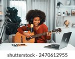 Host channel in musician of young African American playing guitar along with singing, broadcasting on laptop in studio. Decoration of equipment of headsets and recording microphone. Tastemaker.