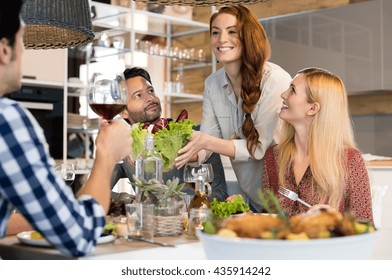 Host Woman Serving Salad To Guests At Home. Smiling Woman Serving Salads To Her Friends At Home. Happy Smiling People Eating Together For Lunch. Group Of Friends Enjoying Meal At Home Together
