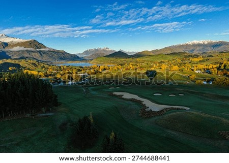 A hot air balloon casts a sharp shadow over a green fairway, with The Remarkables mountain range and Lake Wakatipu visible in the background under crisp autumn morning light.