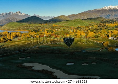 A hot air balloon casts a sharp shadow over a green fairway, with The Remarkables mountain range and Lake Wakatipu visible in the background under crisp autumn morning light.