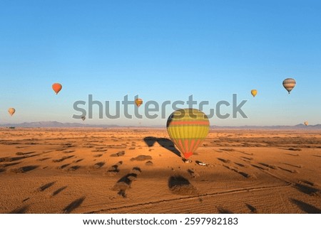 Hot air balloons float over a vast desert landscape at sunrise, casting long shadows on the golden sand. The clear sky and distant mountains add depth.