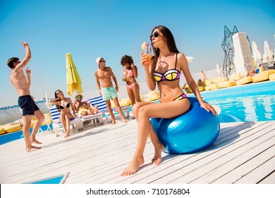 Hot brunette gorgeous girl in sun protective eye wear and bikini, drinking juice by the swim pool, sit on huge baloon, so attractive, ideal, her skin is so perfect and healthy, shining and smooth