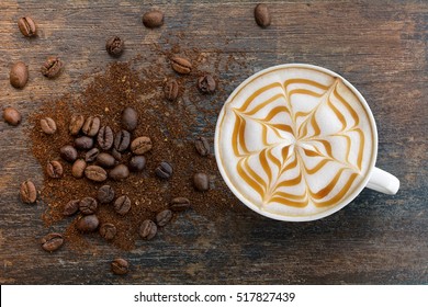 Hot Cappuccino coffee with zigzag caramel motif or spiderweb art floating on top and coffee beans with ground on wooden background. Coffee break at retro style coffee shop,t op view.