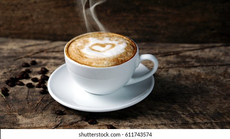 Hot Cappuccino With Streamed Milk. Pour The Hot Milk Made A Heart. A Cup Of Strong Coffee Put On The Wood Table With Shiny Dark Roasted Italian Coffee Beans. Aroma Espresso And Flavor Beverage.