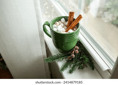 Hot chocolate in a green mug with mini marshmallows and cinnamon sticks. Sitting on a window sill with fairy lights and pine sprig with frosted berries.  - Powered by Shutterstock