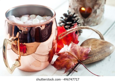 Hot Chocolate, Topped With Marshmallow And Served In A Shiny Copper Mug. On A White Wooden Table With Autumn Leaves, Pine Cone And Wooden Table Decoration.