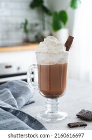 Hot chocolate with whipped cream and chocolate pieces in a tall glass with a cinnamon stick on the background of a white kitchen in the  early morning. Front view and close up.