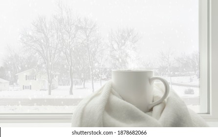 Hot Cocoa Mug Covered By White Comfort Soft Wool Scarf At A Frosted Glass Window In Warm And Cozy Home With Beautiful Landscape Scene Of Snow Falling Outside. White Winter Concept. Cold Weather.