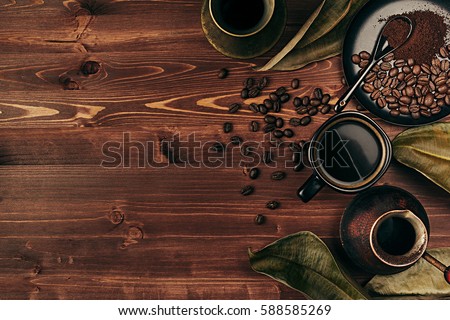 Hot coffee in black cup and several turkish pots cezve with beans, dry leaves with copy space on brown old wooden board background, top view. Rustic style.