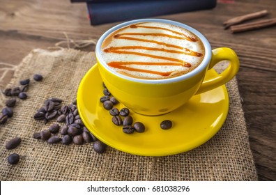 Hot Coffee Caramel  Macchiato In Yellow Cup With Coffee Bean And Sack Cloth On Wooden Table ,notebook With Cinnamon  Stick For Decoration. 