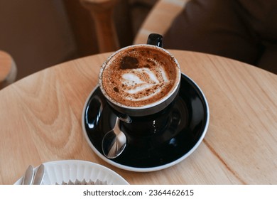 Hot coffee with latte art in black  ceramic cup on wooden table. Cappuccino or latte with frothy foam milk. Cafe and bar, barista art concept. - Shutterstock ID 2364462615