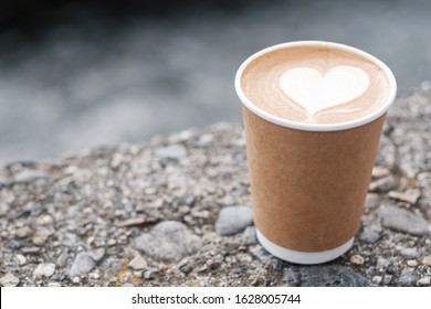 Hot coffee paper cup against river background, heart shape latte coffee art. Love, holiday, Valentine day and free plastic container concept