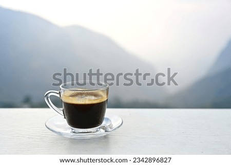 Hot coffee in a transparent glass cup with saucer placed on a white table on a blurred natural background. Concept of drinking coffee in the morning in the calm atmosphere of nature on a long weekend