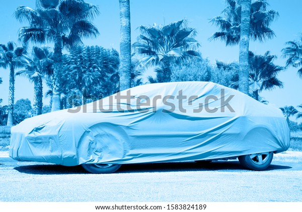 Hot countries, protection from heating\
the car. Parked car covered with a cover from the sun\'s raysin in\
classic blue color ton. The car stands amid\
parlm.