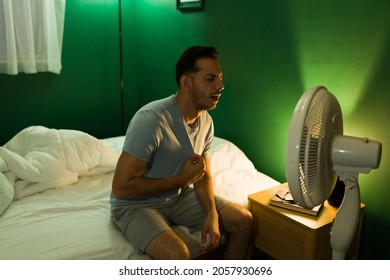 It's So Hot. Distressed Latin Man Sitting In Front Of The Ventilator In Bed And Can't Sleep Because Of The Heat