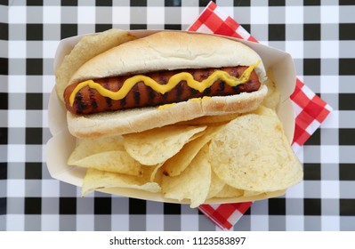 Hot Dog. Hot Dog In Bun And Potato Chips With Mustard Squiggle On A Black And White Checker Serving Tray. Isolated On White. Room For Text. 