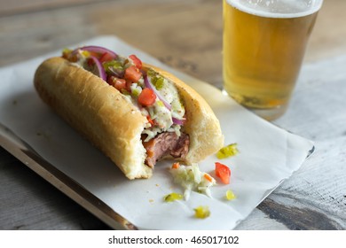 Hot Dog With Coleslaw, Tomato, Onions, Relish And Bite Missing. Shown With Glass Of Cold Beer. 