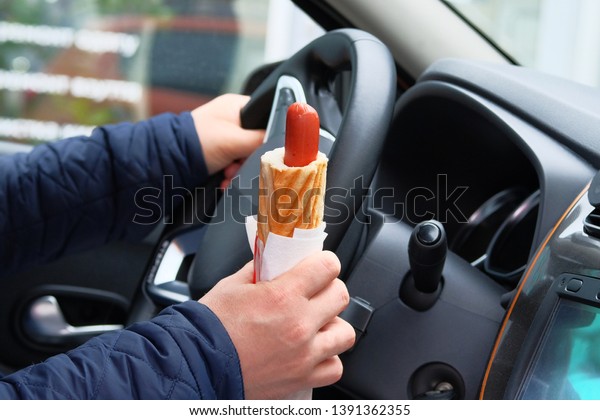 Hot dog in\
driver hand in car on sunny blurred background. Takeaway, social\
media. Snack in man hand in\
auto.